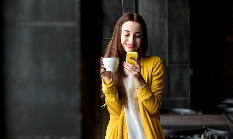 Young and pretty woman in yellow sweater using phone holding a cup of coffee in the dark cafe interior