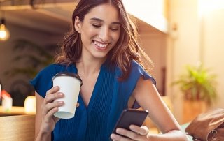 Young beautiful woman holding coffee paper cup and looking at smartphone while sitting at cafeteria. Happy university student using mobile phone. Businesswoman in casual clothes drinking coffee, smiling and using smartphone indoor.