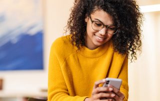 happy woman in eyeglasses using smartphone while sitting at table indoors